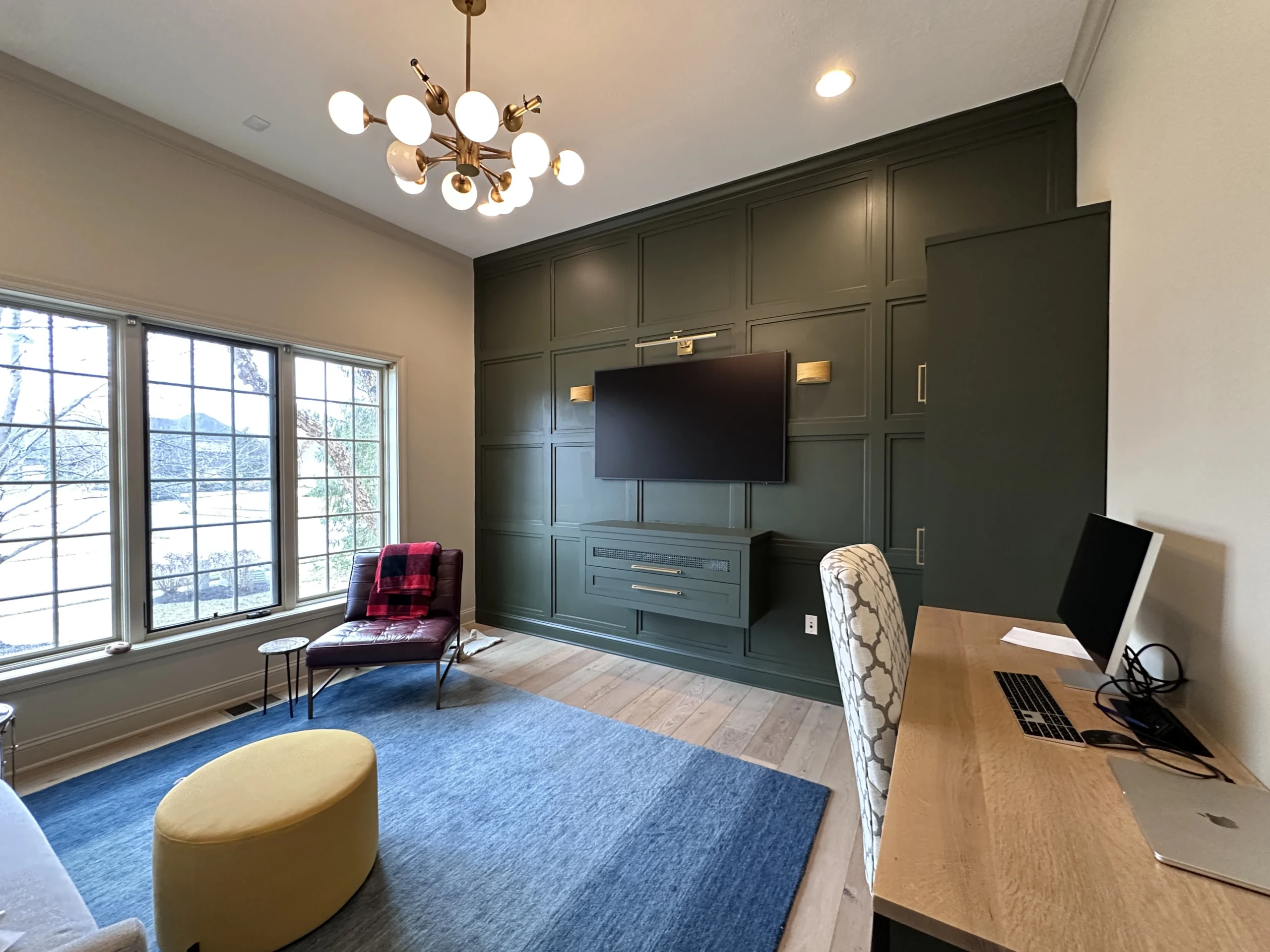Finished den and home office with dark green paneled accent wall, custom cabinetry, desk, seating area, and wood flooring.